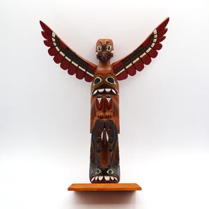 May include: A carved wooden totem pole featuring an eagle with outstretched wings. The totem is primarily brown with red, black, and white accents. It stands on a small, rectangular wooden shelf against a white background.