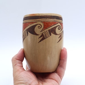 May include: A cylindrical ceramic vessel with a smooth tan base and a decorative band near the rim. The band features a repeating pattern of dark brown spiral wave motifs and geometric shapes set against a burnt orange background.