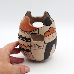 May include: Hand-painted ceramic vessel with a unique design. The beige-colored pot features black, orange, and brown geometric patterns and shapes. The top has two small figures. The vessel has a hole in the middle.