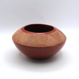May include: A round, terracotta-colored ceramic bowl with a smooth, dark red finish on the lower half and a wide, textured band around the upper shoulder. The textured section features a repeating pattern of incised, diagonal parallel lines.