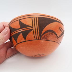 May include: Handmade orange ceramic bowl with black geometric designs. The bowl features a rounded shape and is adorned with intricate patterns, including triangles and lines. The design is symmetrical and appears to be hand-painted, showcasing a traditional craft.