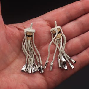 May include: Two handmade earrings with a unique design. Each earring features a woven basket-like top with black and gold accents, long white cords, and silver-colored metal cone-shaped dangles. The earrings are displayed on a neutral background.