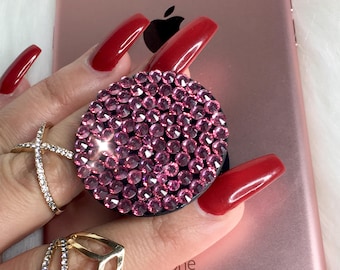 May include: Rose gold smartphone with a pink rhinestone phone grip. The phone grip is round and covered in small, sparkling pink rhinestones. The background is a soft, white, fluffy texture.