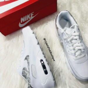 May include: White leather sneakers with a white swoosh logo and a row of clear rhinestones around the edges. The sneakers are on a white fuzzy surface. The text "@VioletsPinkBoutique" is visible in the image.