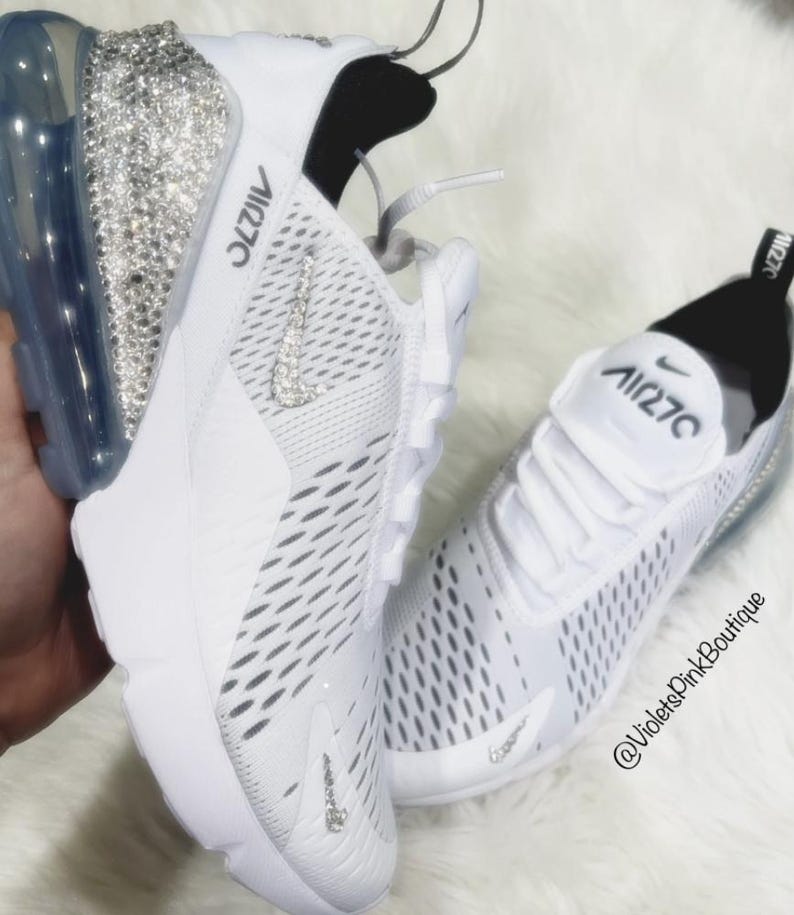 Puede incluir: Zapatillas deportivas blancas con el texto "Air 270" en la leng&uuml;eta y el lateral. El tal&oacute;n de una zapatilla est&aacute; cubierto de brillantes pedrer&iacute;a. El logo de Nike swoosh tambi&eacute;n est&aacute; adornado con pedrer&iacute;a. Las zapatillas est&aacute;n sobre una superficie blanca y esponjosa.