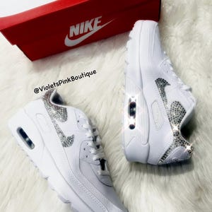 May include: White leather Nike Air Max 90 sneakers with a silver rhinestone swoosh and a white Nike box. The text '@VioletsPinkBoutique' is visible on the box.