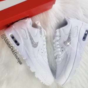 May include: Pair of white sneakers with a sparkling silver Nike swoosh. The shoes are displayed on a fluffy white surface, with a red box in the background. The text "@VioletsPinkBoutique" is visible.