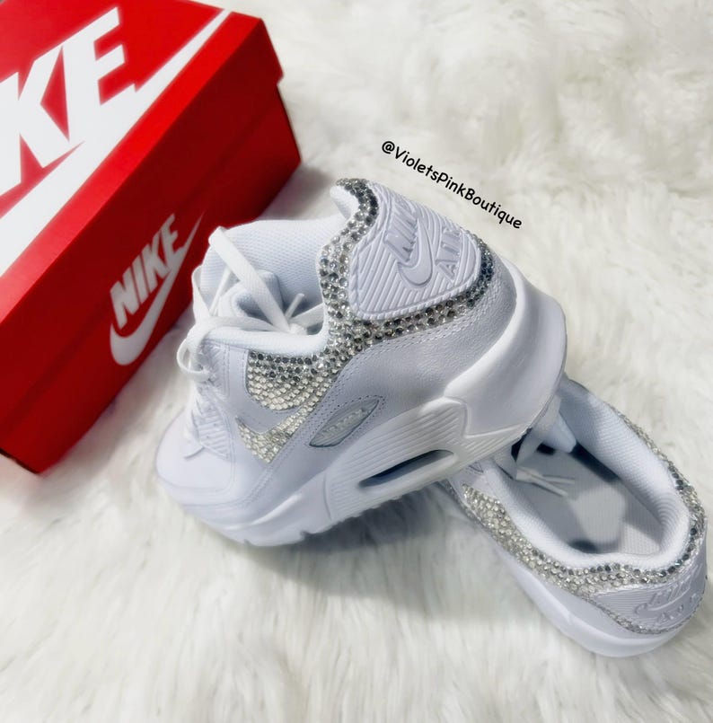 May include: White Nike Air Max 90 sneakers with a white swoosh logo and a bedazzled design. The sneakers are on a white fluffy surface. The text "@VioletsPinkBoutique" is visible in the image.