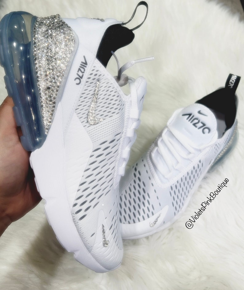 Puede incluir: Zapatillas deportivas blancas con el texto "AIR 270" en negro. Las zapatillas tienen una suela transparente de burbujas de aire y est&aacute;n adornadas con pedrer&iacute;a. El logotipo de Nike swoosh tambi&eacute;n est&aacute; adornado con pedrer&iacute;a. Las zapatillas est&aacute;n sobre una superficie blanca y esponjosa.