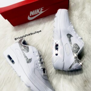 May include: White leather Nike Air Max 90 sneakers with a silver rhinestone swoosh and a white Nike box. The text '@VioletsPinkBoutique' is visible on the box.