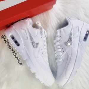 May include: Pair of white sneakers with a sparkling silver Nike swoosh. The shoes are displayed on a fluffy white surface, with a red box in the background. The text "@VioletsPinkBoutique" is visible.