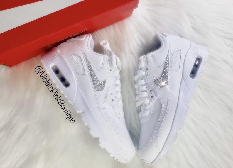 May include: Pair of white sneakers with a sparkling silver Nike swoosh. The shoes are displayed on a fluffy white surface, with a red box in the background. The text "@VioletsPinkBoutique" is visible.