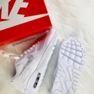 May include: White Nike Air Max 90 sneakers with crystal embellishments. The sneakers are displayed with a red Nike box on a white fluffy surface. The sole of one shoe is visible, and the other shoe is on its side.