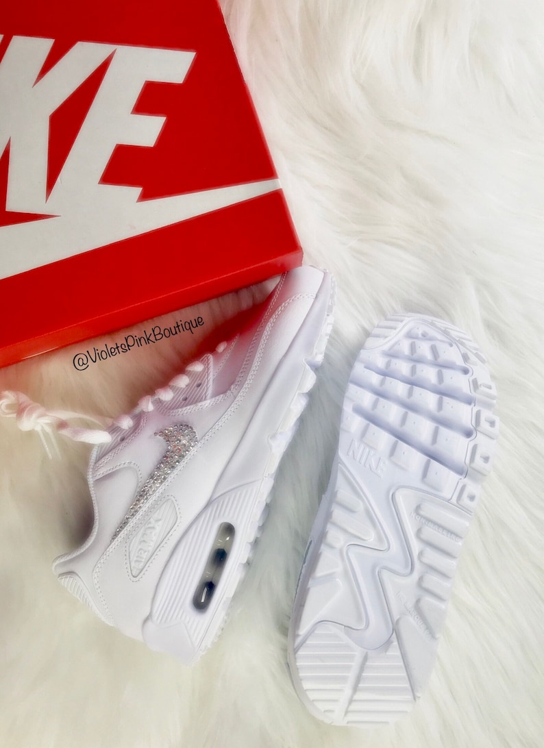 May include: White Nike Air Max 90 sneakers with crystal embellishments. The sneakers are displayed with a red Nike box on a white fluffy surface. The sole of one shoe is visible, and the other shoe is on its side.