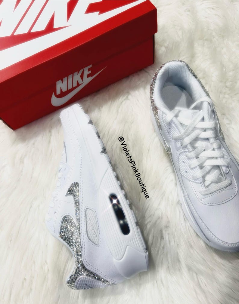 May include: White leather sneakers with a white swoosh logo and a row of clear rhinestones around the sole. The sneakers are on a white fuzzy surface. The text "@VioletsPinkBoutique" is visible in the image.