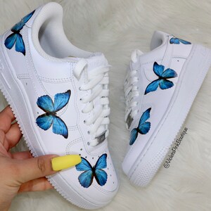 May include: White low-top sneakers with blue butterfly designs painted on them. The butterflies are different sizes and are painted on the sides and toes of the shoes. The shoes are tied with white laces and have a white sole.