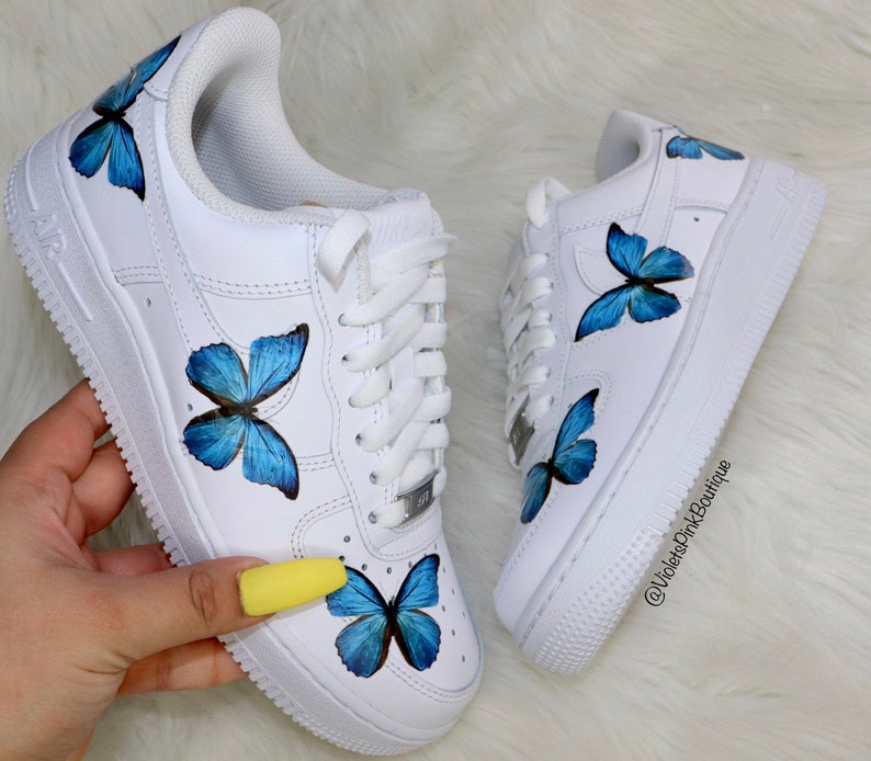 May include: White low-top sneakers with blue butterfly designs painted on them. The butterflies are different sizes and are painted on the sides and toes of the shoes. The shoes are tied with white laces and have a white sole.