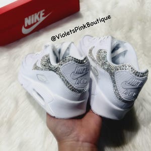 May include: White leather Nike Air Max 90 sneakers with a crystal embellishment on the swoosh and toe. The sneakers are on a white furry surface. The text "@VioletsPinkBoutique" is visible on the image.