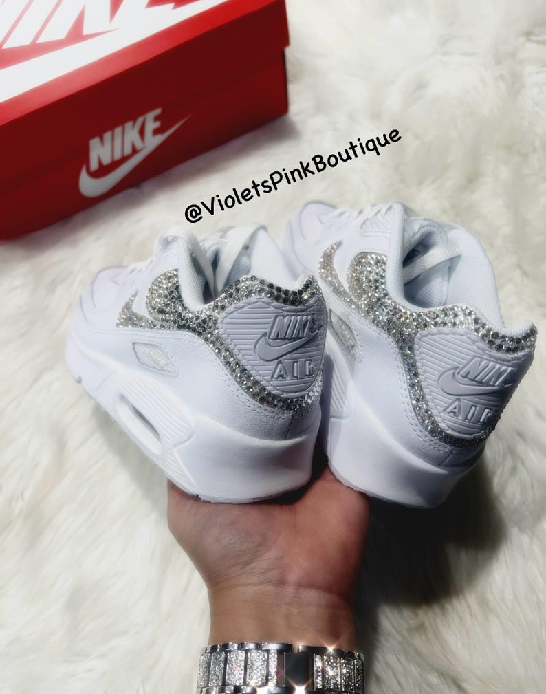May include: White leather Nike Air Max 90 sneakers with a crystal embellishment on the swoosh and toe. The sneakers are on a white furry surface. The text "@VioletsPinkBoutique" is visible on the image.