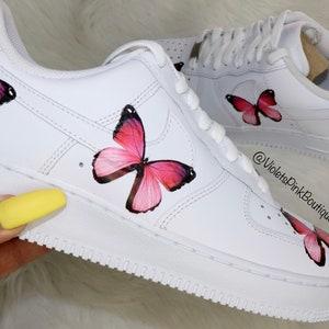 May include: White leather sneakers with pink butterfly designs painted on them. The sneakers have a white swoosh logo and the text "AIR" on the side. The text "@VioletsPinkBoutique" is written on the side of the shoe.