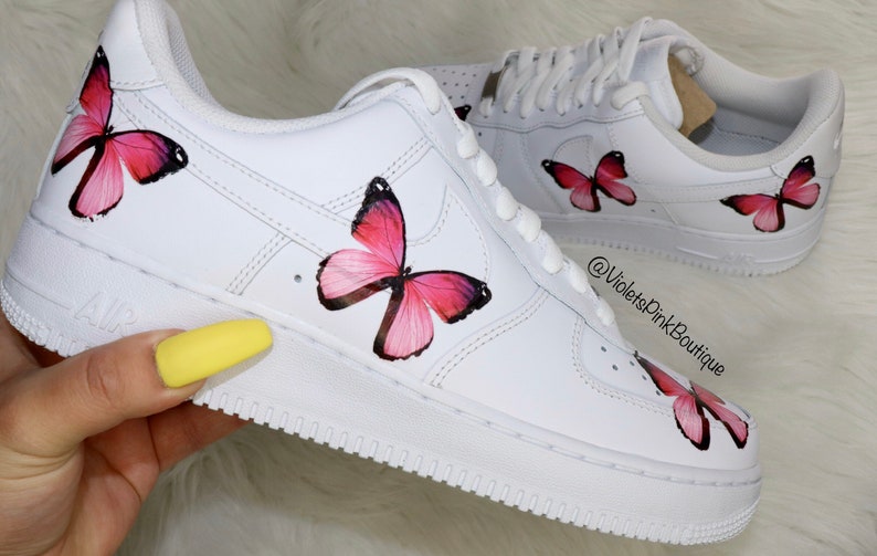 May include: White leather sneakers with pink butterfly designs painted on them. The sneakers have a white swoosh logo and the text "AIR" on the side. The text "@VioletsPinkBoutique" is written on the side of the shoe.
