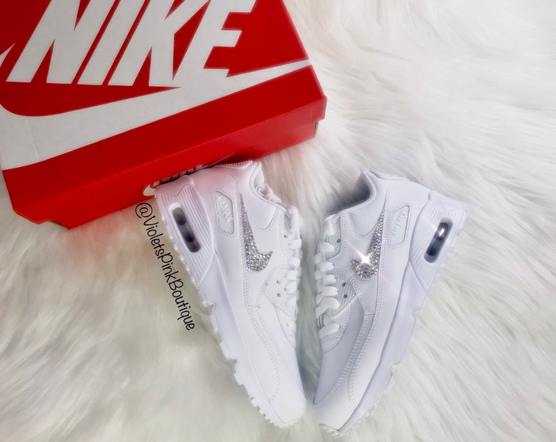 May include: White Nike Air Max 90 sneakers with crystal embellishments on the Nike swoosh logo. The shoes are displayed on a white fluffy surface next to a red Nike box with a white logo.