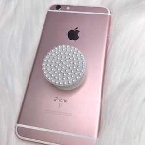 Puede incluir: iPhone S color oro rosa con un soporte para teléfono blanco con perlas. El soporte para teléfono tiene el texto "POPCRETS.COM" impreso. El teléfono está sobre una superficie blanca y esponjosa.