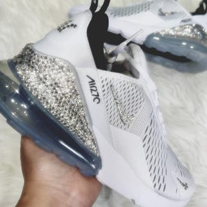 Puede incluir: Zapatillas blancas Nike Air Max 270 con adornos de cristal en el tal&oacute;n y el logotipo de Nike. Las zapatillas tienen una parte superior blanca con una leng&uuml;eta negra y una suela de burbuja de aire transparente. El texto "Air 270" es visible en el lateral.