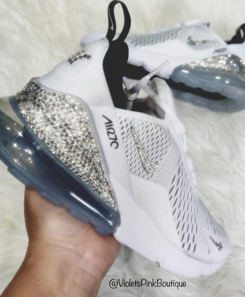 Puede incluir: Zapatillas blancas Nike Air Max 270 con adornos de cristal en el tal&oacute;n y el logotipo de Nike. Las zapatillas tienen una parte superior blanca con una leng&uuml;eta negra y una suela de burbuja de aire transparente. El texto "Air 270" es visible en el lateral.