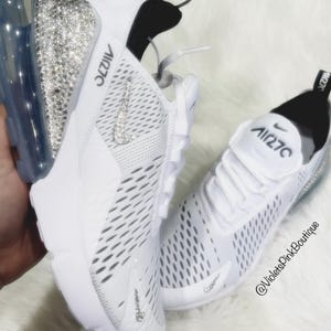 Puede incluir: Zapatillas deportivas blancas con el texto "Air 270" en la leng&uuml;eta y el lateral. El tal&oacute;n de una zapatilla est&aacute; cubierto de brillantes pedrer&iacute;a. El logo de Nike swoosh tambi&eacute;n est&aacute; adornado con pedrer&iacute;a. Las zapatillas est&aacute;n sobre una superficie blanca y esponjosa.