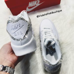 May include: A pair of white leather sneakers with rhinestones on the sides and the Nike logo on the back. The sneakers are on a white fuzzy surface. The text '@VioletsPinkBoutique' is on the image.