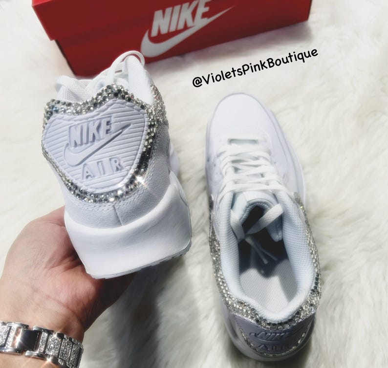 May include: A pair of white leather sneakers with rhinestones on the sides and the Nike logo on the back. The sneakers are on a white fuzzy surface. The text '@VioletsPinkBoutique' is on the image.