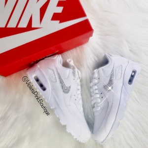 May include: White Nike Air Max 90 sneakers with crystal embellishments on the Nike swoosh logo. The shoes are displayed on a white, fluffy surface next to a red Nike shoe box.