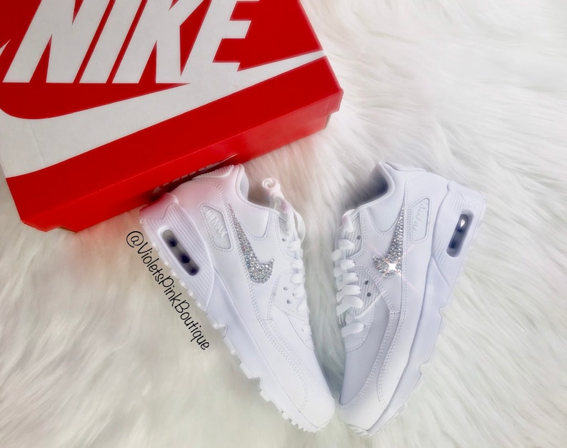 May include: White Nike Air Max 90 sneakers with crystal embellishments on the Nike swoosh logo. The shoes are displayed on a white, fluffy surface next to a red Nike shoe box.