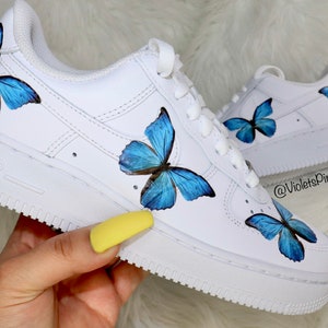 May include: White leather low-top sneakers with blue butterfly designs painted on them. The sneakers have a white sole with the word "AIR" printed on the side. The text "@VioletsPinkBoutique" is printed on the side of the shoe.