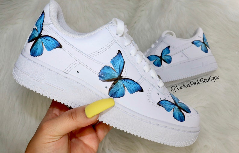 May include: White leather low-top sneakers with blue butterfly designs painted on them. The sneakers have a white sole with the word "AIR" printed on the side. The text "@VioletsPinkBoutique" is printed on the side of the shoe.