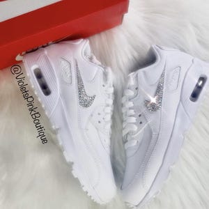 May include: Pair of white sneakers with crystal embellishments on the Nike swoosh logo. The shoes are displayed on a white fluffy surface, with a red and white box in the background. The text "@VioletsPinkBoutique" is visible.