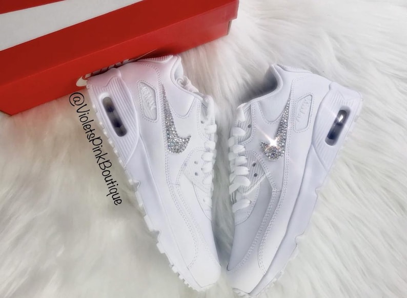 May include: Pair of white sneakers with crystal embellishments on the Nike swoosh logo. The shoes are displayed on a white fluffy surface, with a red and white box in the background. The text "@VioletsPinkBoutique" is visible.