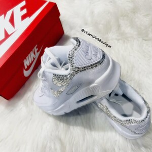May include: White Nike Air Max 90 sneakers with a white swoosh logo and a bedazzled design. The sneakers are on a white fluffy surface. The text "@VioletsPinkBoutique" is visible in the image.