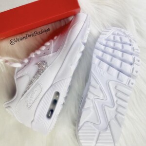 May include: White Nike Air Max 90 sneakers with a sparkling silver Nike logo. The sneakers are displayed with the sole of one shoe visible, and a red box is in the background. The text "@VioletsPink Boutique" is visible.