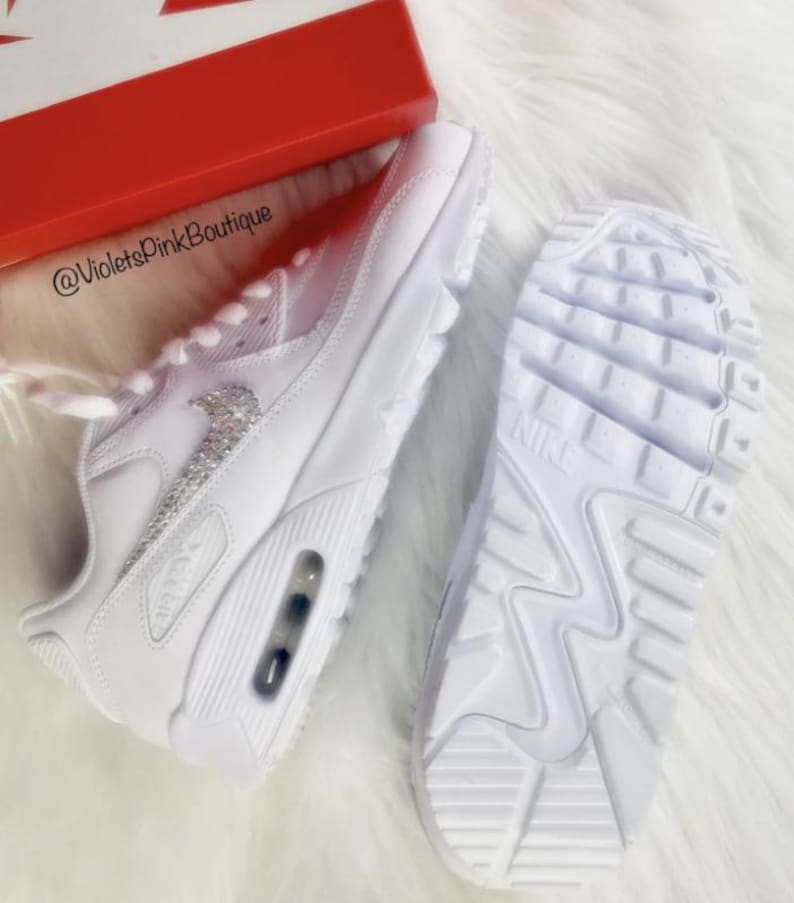 May include: White Nike Air Max 90 sneakers with a sparkling silver Nike logo. The sneakers are displayed with the sole of one shoe visible, and a red box is in the background. The text "@VioletsPink Boutique" is visible.