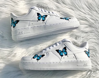 May include: White leather low-top sneakers with blue butterfly designs painted on them. The sneakers have the word "AIR" printed on the side. The text "@VioletsPinkBoutique" is visible in the top right corner of the image.