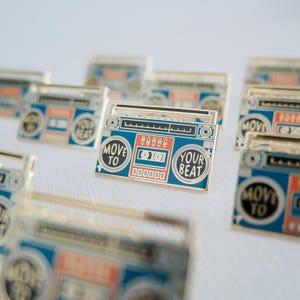 May include: Enamel pins in the shape of a retro boombox. The pins are blue, red, and gold with the words "Move to Your Beat". The pins are arranged on a white surface.