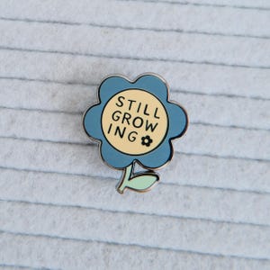 May include: A blue flower-shaped enamel pin with the words "STILL GROWING" in black text on a yellow center. The pin has a small black flower graphic and a green leaf.