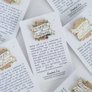May include: Gold enamel pin with a white banner that reads "Strength in Softness". The banner is decorated with pink and green flowers. The pin is on a white card with text that reads "Being soft is not a weakness; it takes courage to embrace your vulnerability. Feel it all and be kind to yourself in the process. Allow yourself to fall apart and then gather yourself back up with strength. Dare to be real and not let the fear of judgment hold you back. Stay soft in this tough world; in your gentleness, you'll find strength, compassion and connection. Enamel Pin THEWILDARTSCAPADE.COM DESIGNED IN SINGAPORE"