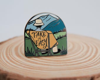 May include: Enamel pin featuring a person in a folding chair with the text "Take it Easy." The pin has a mountain and landscape background with green, blue, and brown colors. The person is wearing a hat and a yellow blanket.