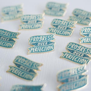 May include: A collection of teal blue enamel pins with gold accents. Each pin features a banner with the text "Progress Over Perfection".