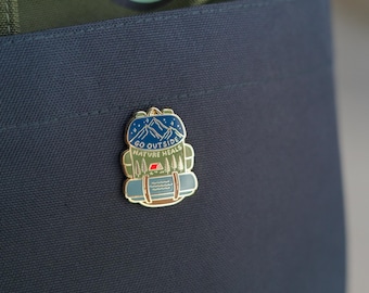 May include: A gold enamel pin with a blue and green backpack design. The backpack has a mountain scene with stars and the text "GO OUTSIDE NATURE HEALS".