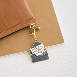 May include: A gold-coloured charm with a blue envelope design, attached to a brown wallet. The envelope charm has the text "my Best is always good enough" on a white card, with leaf and heart details.
