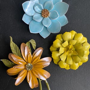 May include: Three floral brooches featuring layered petals. One is light blue with a textured gold center, one is bright yellow, and one is orange with green leaves and a central clear rhinestone. Each piece displays a glossy, painted finish.